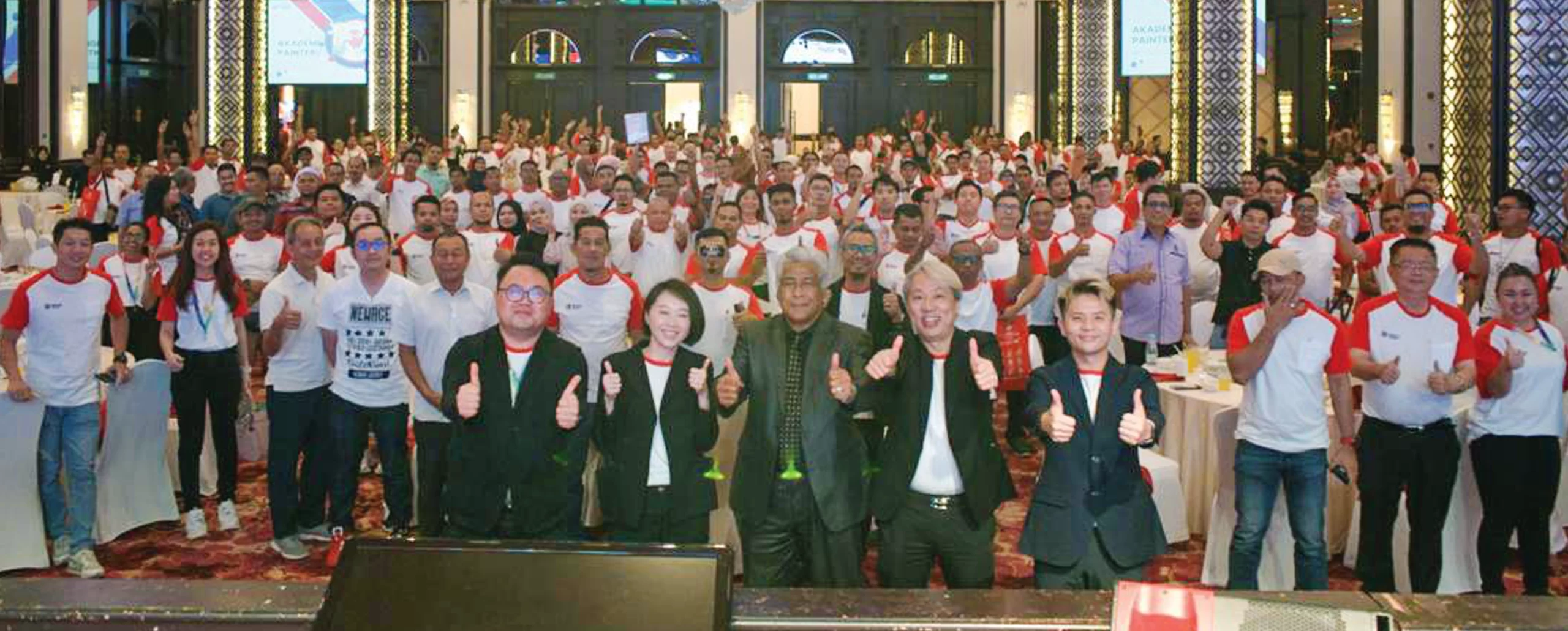 Group photo of attendees at a Nippon Paint event, showcasing teamwork and enthusiasm, with participants wearing branded shirts and giving thumbs up in a celebratory atmosphere.