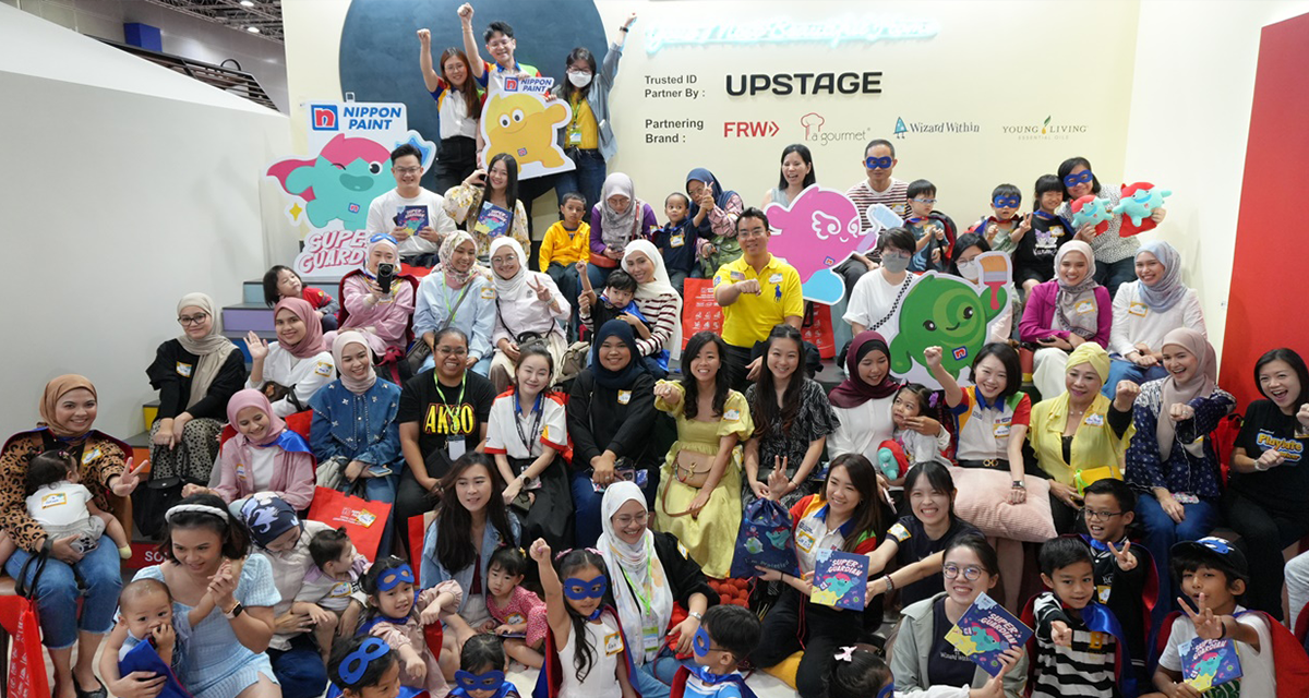 A large group of diverse participants, including families and children, gather at a Nippon Paint event, showcasing colorful promotional materials and engaging activities. The backdrop features branding and logos of Nippon Paint and partnering brands, highlighting community involvement and creativity.