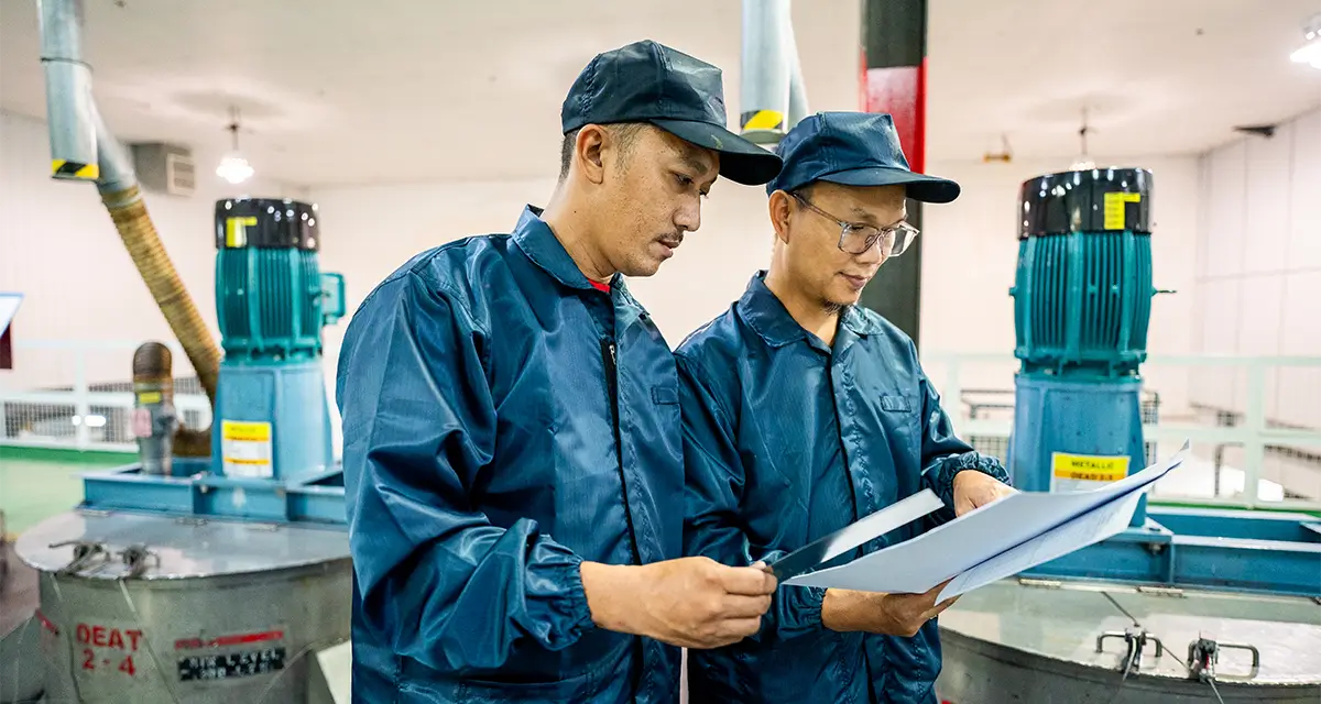 Two workers in blue uniforms and caps review documents in a Nippon Paint production facility, surrounded by industrial machinery.