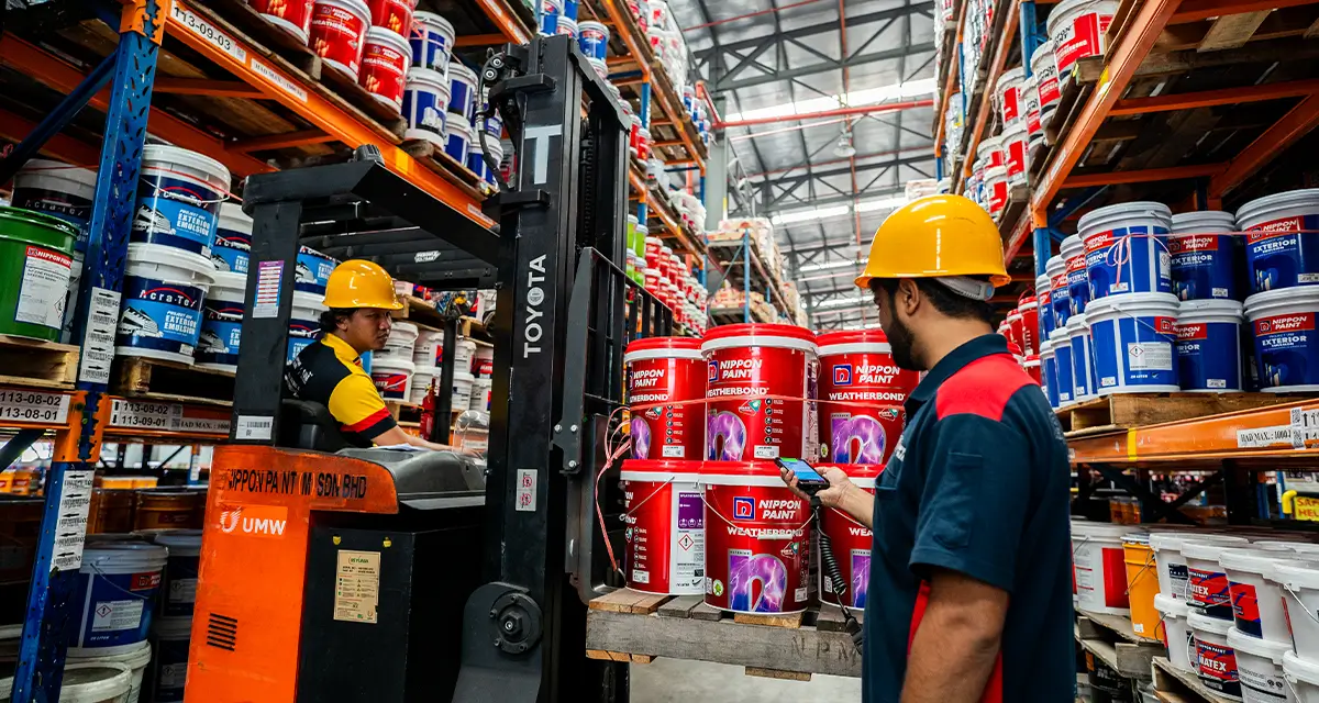 Workers in a Nippon Paint warehouse manage inventory using a forklift, surrounded by various paint products, including Nippon Paint Weathbond. Safety helmets are worn for protection in the busy storage environment.