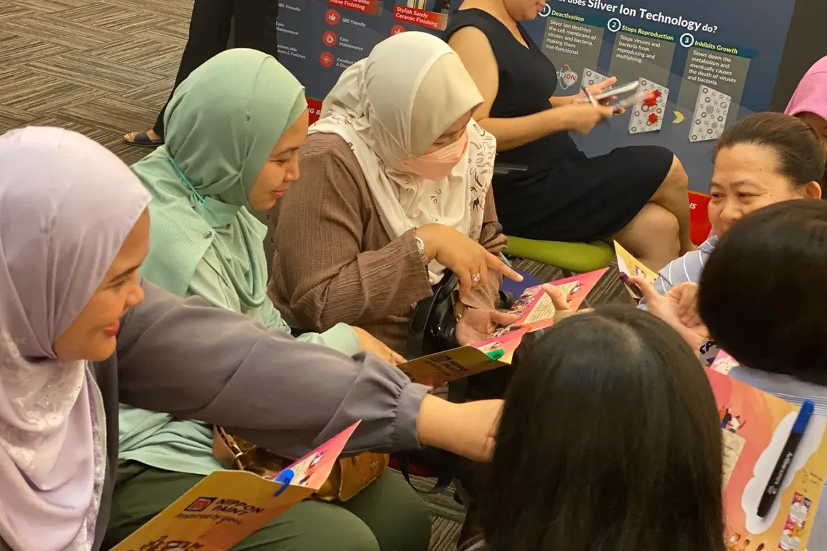 Group of women engaged in a discussion about Nippon Paint products at a promotional event, showcasing color samples and brochures. The setting highlights interactive learning about innovative paint solutions.