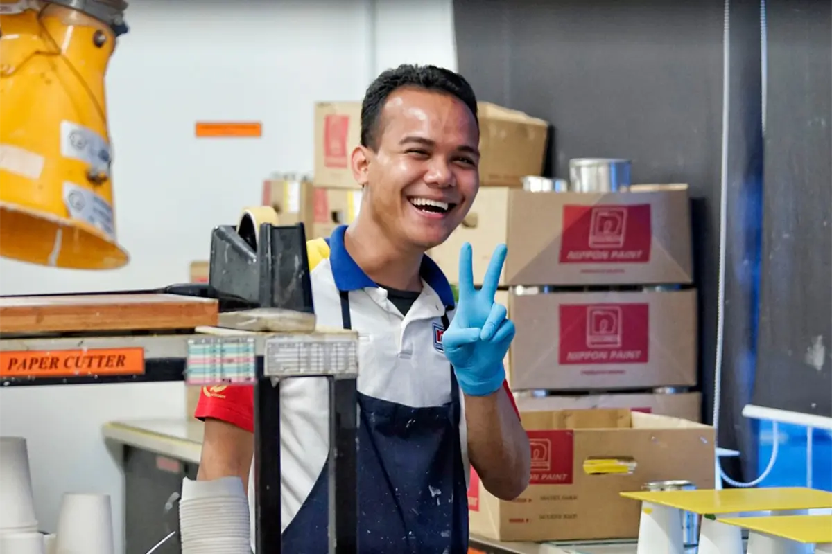 Smiling employee at Nippon Paint workshop, wearing gloves and making a peace sign, surrounded by paint supplies and product boxes.