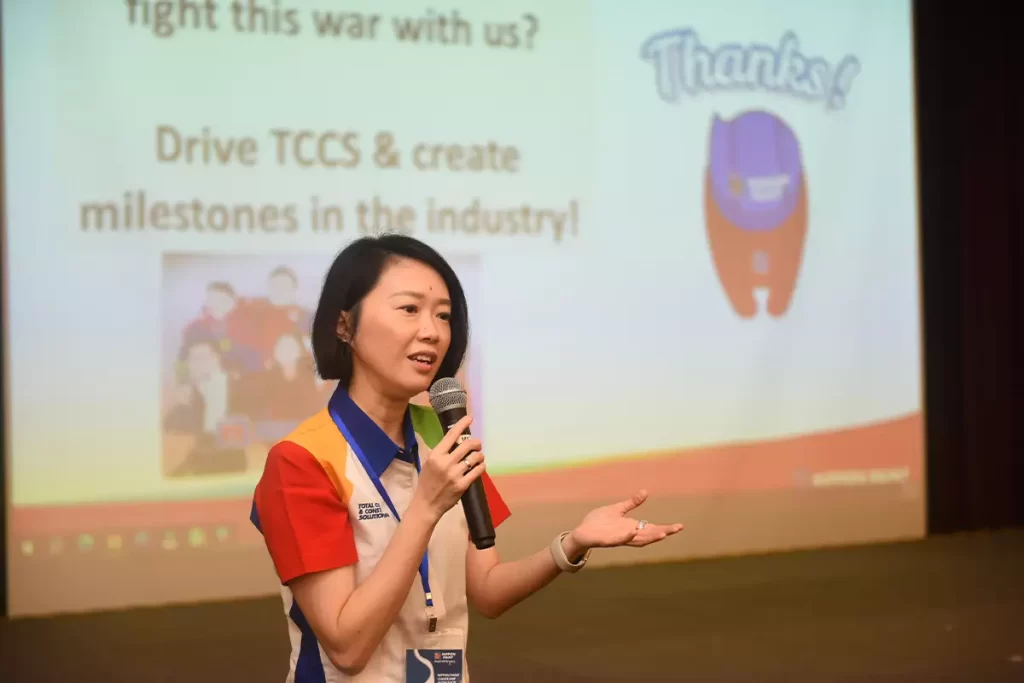 A speaker presenting at a Nippon Paint event, engaging the audience with a microphone and emphasizing the importance of driving TCCS and creating industry milestones, with a colorful backdrop displaying supportive messaging.