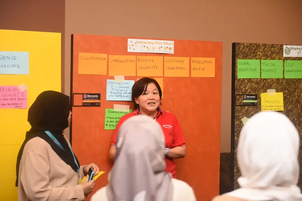 A woman in a red Nippon Paint shirt leads a workshop on communication and collaboration, standing in front of colorful sticky notes outlining key topics. Participants engage in discussion, highlighting the importance of building inclusivity and customer orientation in team dynamics.