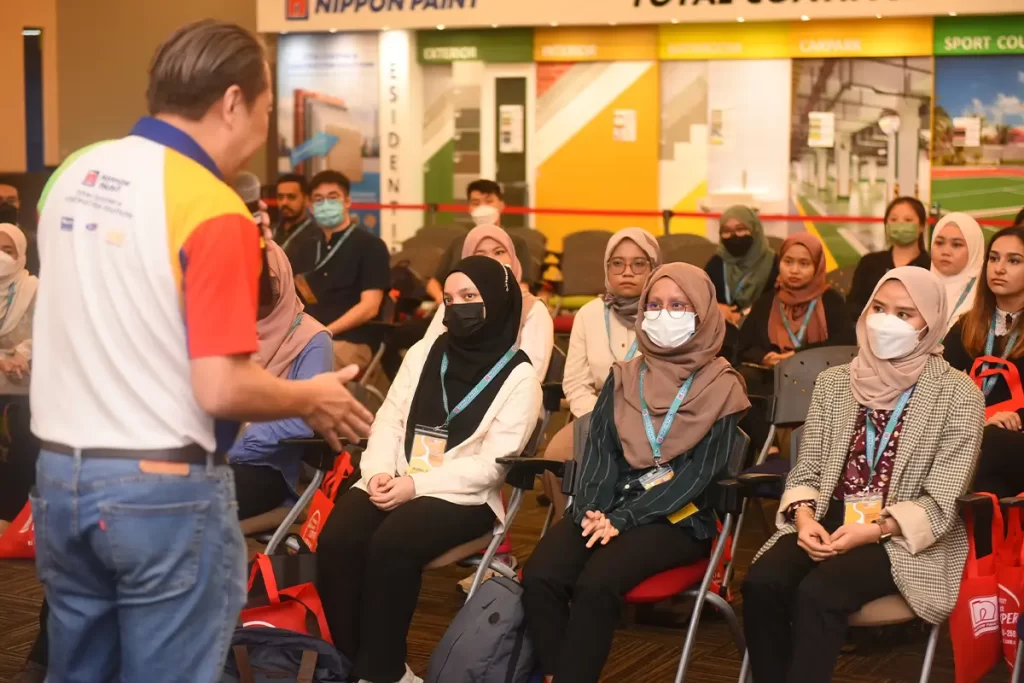 Audience engaged in a presentation at a Nippon Paint event, featuring a speaker addressing a group of attendees wearing masks. The backdrop showcases various Nippon Paint products and branding, highlighting the company's commitment to innovation in the paint industry.