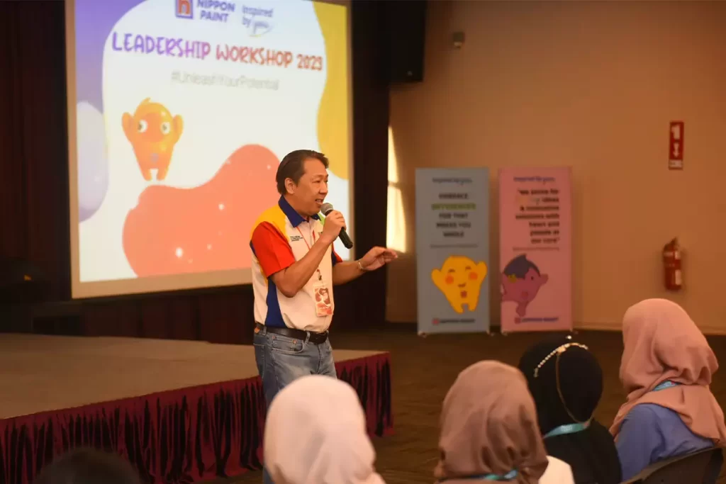 Speaker presenting at the Nippon Paint Leadership Workshop 2023, engaging the audience while emphasizing the theme #UnleashYourPotential, with colorful banners in the background promoting creativity and inspiration.