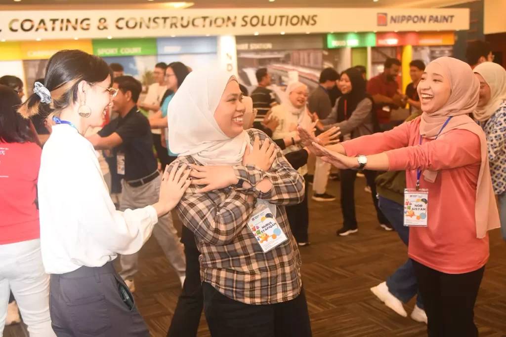 Group of diverse participants engaging in a lively interactive session at a Nippon Paint event, showcasing teamwork and camaraderie. The background features branding and product displays relevant to the coating and construction industry.