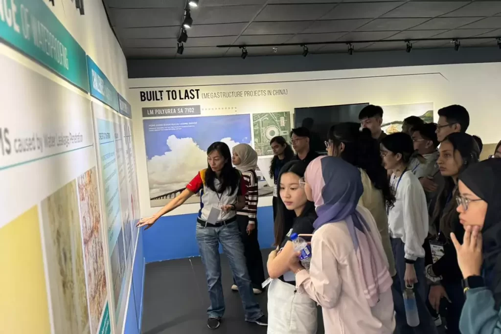 A diverse group of attendees at a presentation, engaging with a display on megastructure projects in China, featuring Nippon Paint products. The presenter points to informative panels while participants listen attentively, highlighting the intersection of architecture and innovative materials.