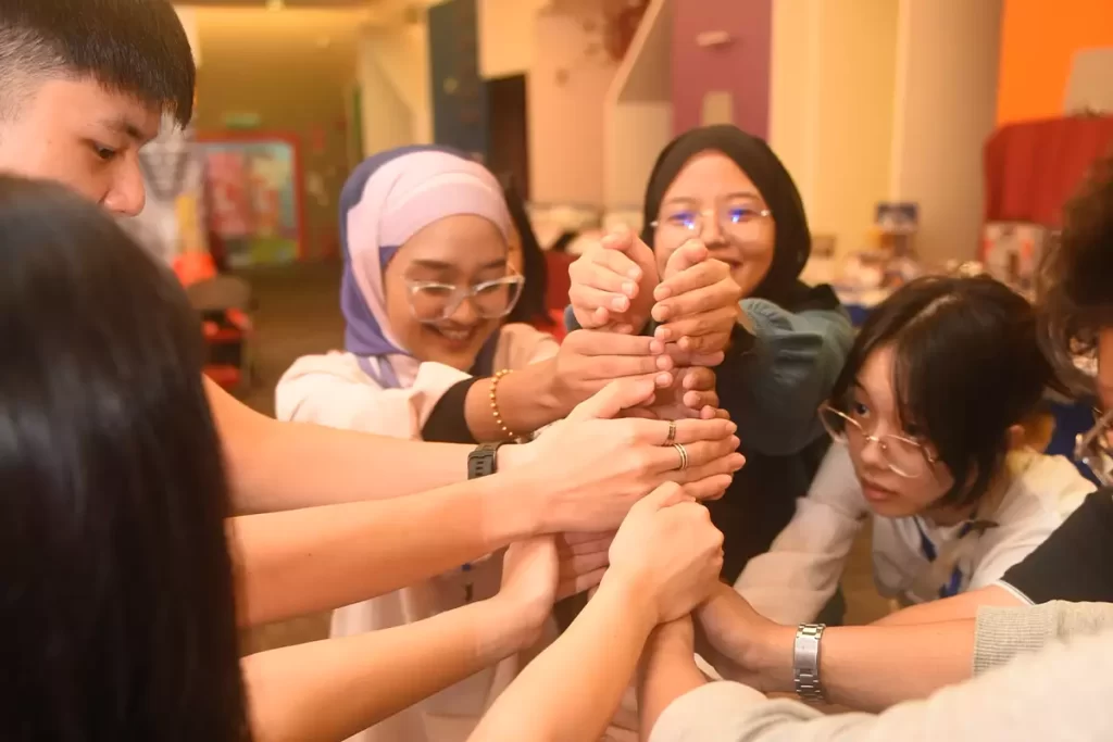 A group of diverse individuals collaboratively stacking their hands together in a team-building activity, showcasing unity and teamwork. The vibrant setting reflects a community event, possibly sponsored by Nippon Paint, emphasizing creativity and collaboration.