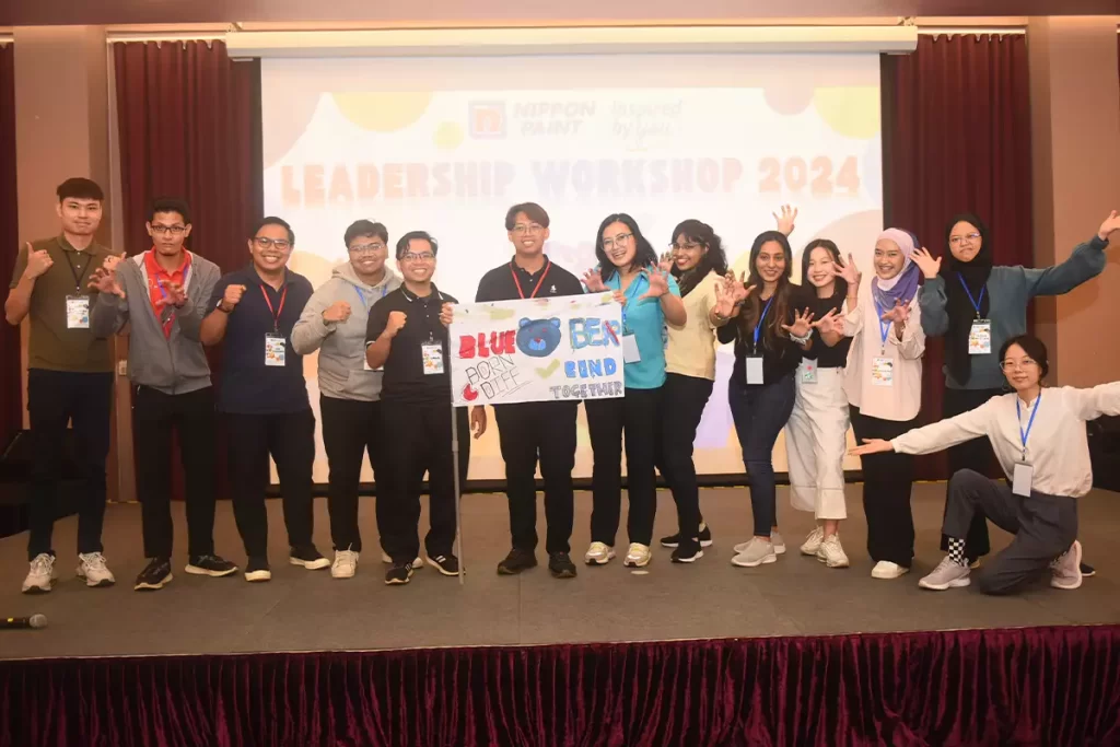 Group photo of participants at the Nippon Paint Leadership Workshop 2024, showcasing teamwork and enthusiasm with a banner that reads "Born Different, Bond Together." The event highlights collaboration and leadership skills among diverse individuals.