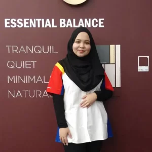 A woman wearing a colorful shirt stands in front of a wall displaying Nippon Paint's "Essential Balance" color palette, featuring the words "Tranquil," "Quiet," "Minimal," and "Natural."
