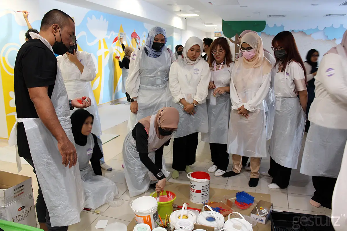 Participants engaged in a creative painting workshop, using Nippon Paint products, while dressed in protective aprons. The scene showcases a vibrant mural in progress, with various paint containers and tools scattered around.