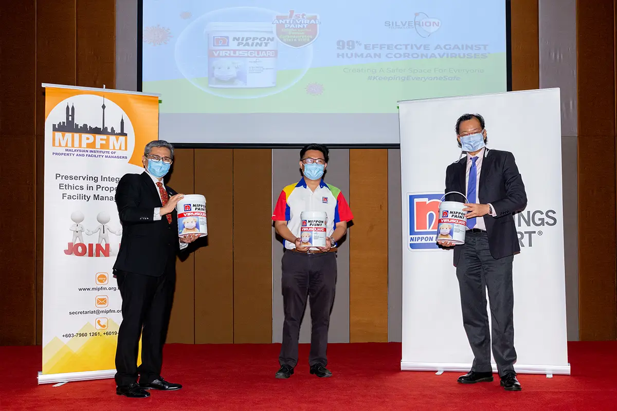 Three professionals hold Nippon Paint's VIRUSGUARD anti-viral paint during a presentation at an event organized by the Malaysian Institute of Property and Facility Managers (MIPFM). The background features a digital screen highlighting the paint's effectiveness against human coronaviruses.