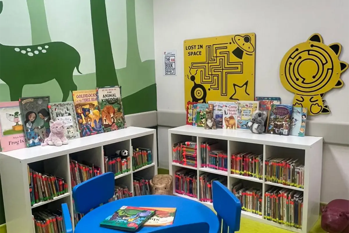 Colorful children's reading area featuring bookshelves filled with various children's books, plush toys, and a blue circular table. The walls are adorned with vibrant murals, including a green deer and playful yellow mazes, all painted with Nippon Paint products to create an inviting and stimulating environment for young readers.