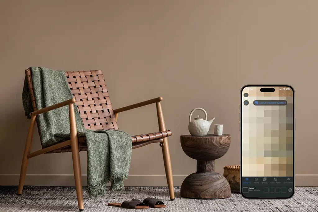Cozy living space featuring a stylish woven chair with a green throw, a wooden side table, and decorative items, complemented by a Nippon Paint color palette app on a smartphone, showcasing modern interior design ideas.