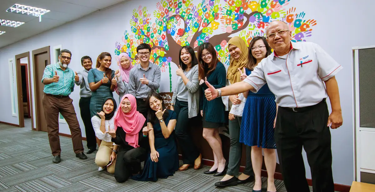 Group of diverse employees posing cheerfully in an office setting, showcasing a colorful hand-painted tree mural featuring Nippon Paint products, symbolizing teamwork and creativity.