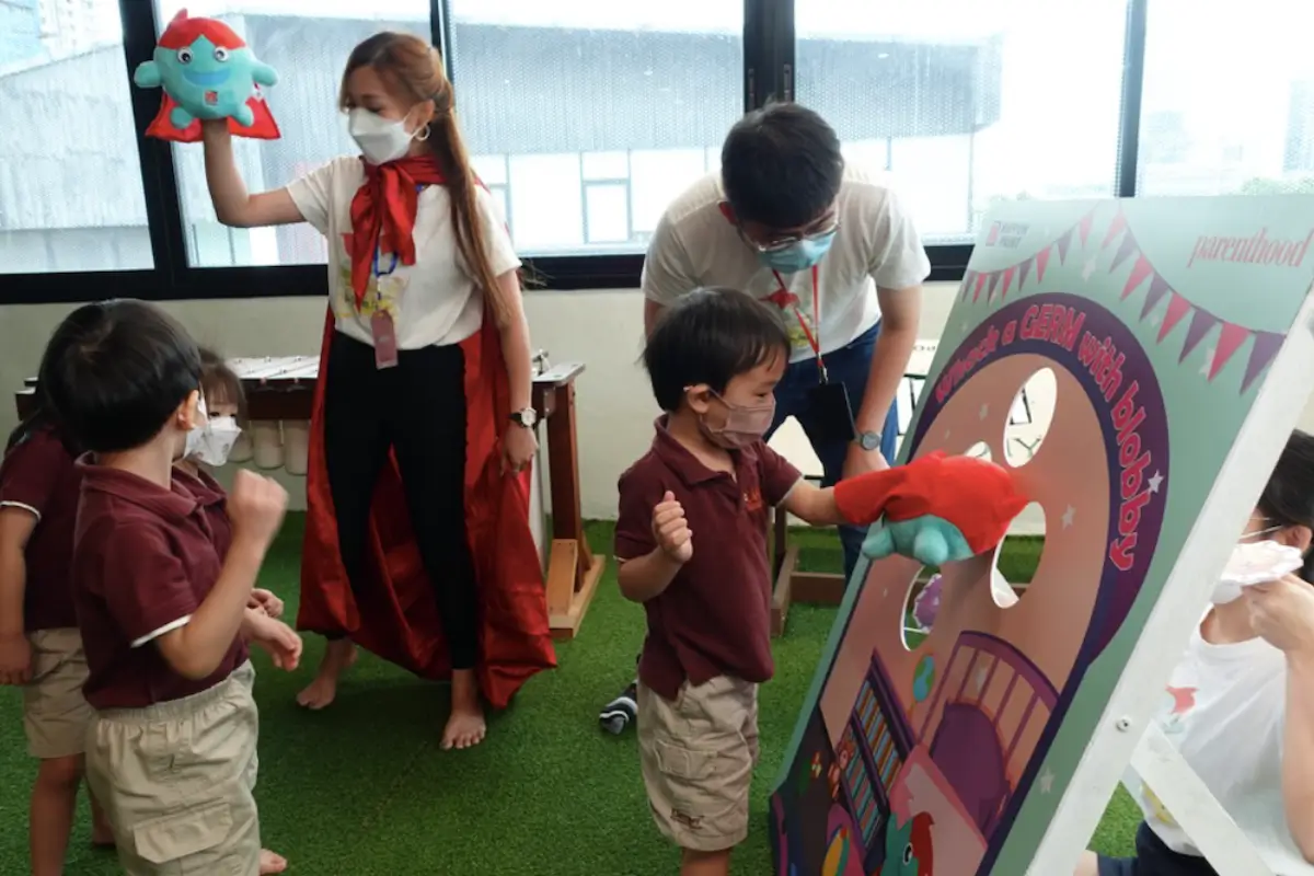 Children engaging in a fun educational activity at a Nippon Paint event, featuring a game to learn about germs with a character named Blobby. The scene shows a teacher in a red cape and mask, guiding students while they interact with colorful props.