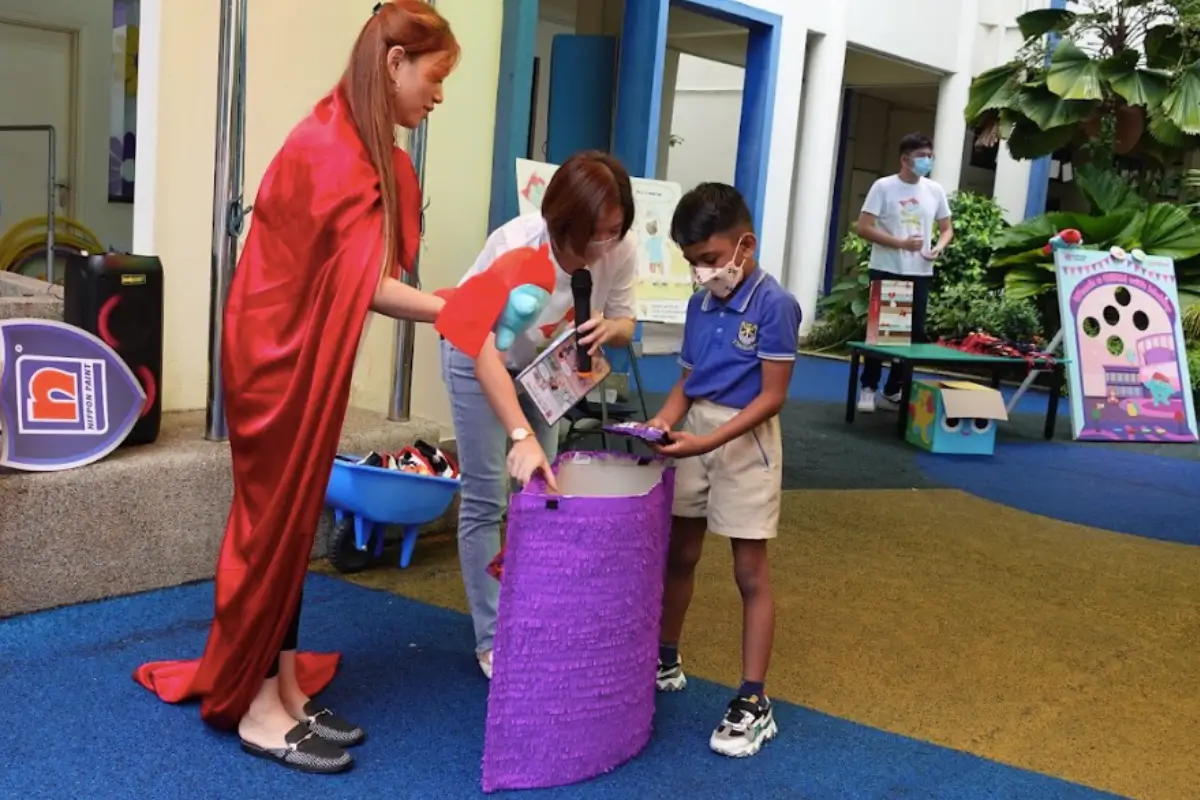 Children participating in a creative workshop, engaging with a purple recycling bin, while two adults facilitate the activity. The Nippon Paint logo is visible in the background, promoting eco-friendly practices and community involvement.