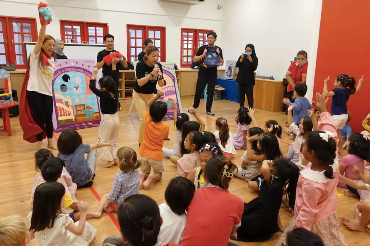 Children engaging in a fun and interactive storytelling session, led by enthusiastic presenters, in a brightly colored classroom. The event promotes creativity and imagination, featuring Nippon Paint branding for a vibrant, inspiring environment.