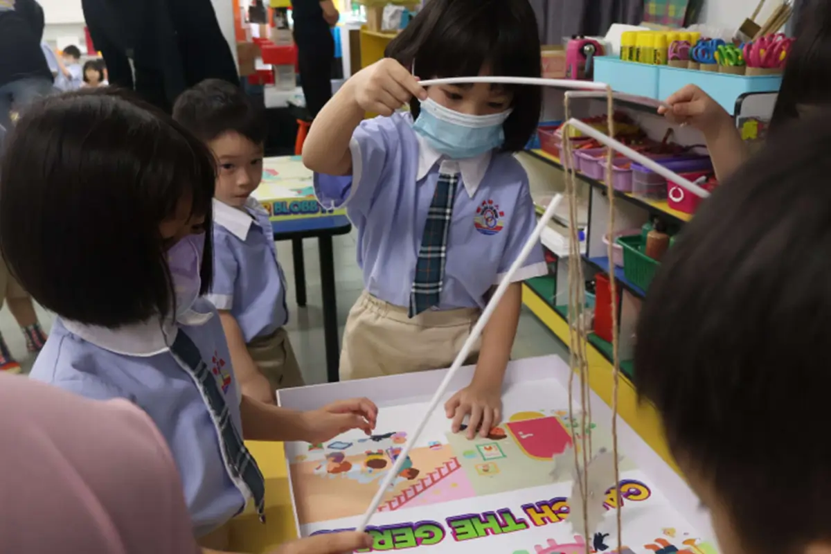 Children engaging in a creative classroom activity, using materials to build a model while wearing masks. The vibrant environment showcases educational tools and colorful Nippon Paint branding, promoting creativity and learning.