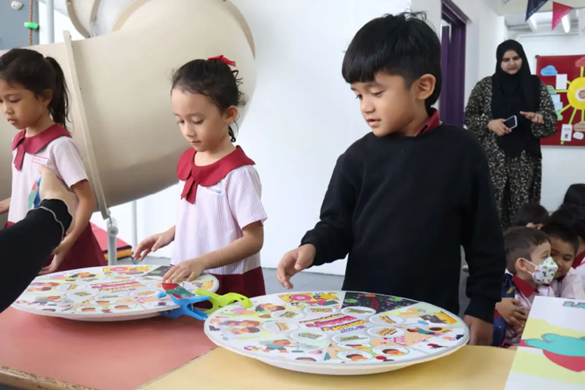 Children engaging in creative activities in a colorful classroom, using Nippon Paint-branded art materials. Two kids focus on their projects while a teacher assists, fostering a vibrant learning environment.