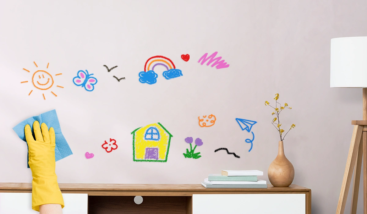 A person cleaning colorful crayon drawings from a wall with a Nippon Paint finish, showcasing a sunny scene with a house, butterflies, and a rainbow.